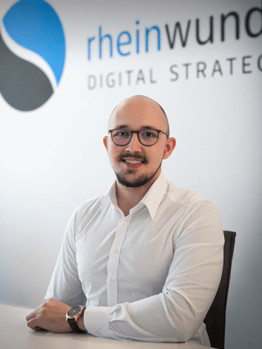 Ein Mann mit Brille sitzt lächelnd an einem Tisch vor einem Logo mit der Aufschrift rheinwunder DIGITAL STRATEGY.