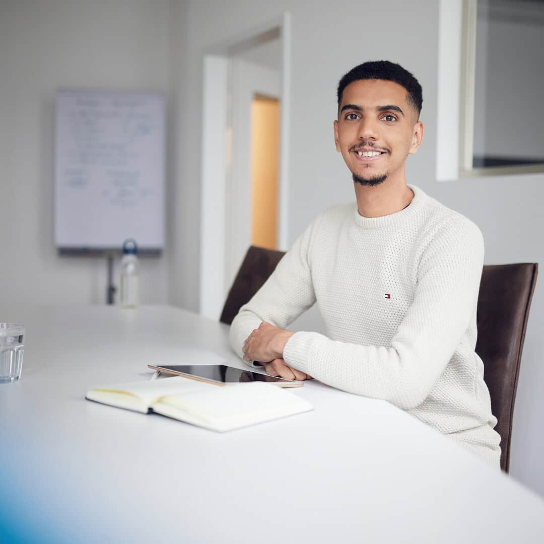 Ein Mann in weißem Pullover sitzt an einem Tisch mit Notizbuch und Tablet. Im Hintergrund ein Whiteboard.