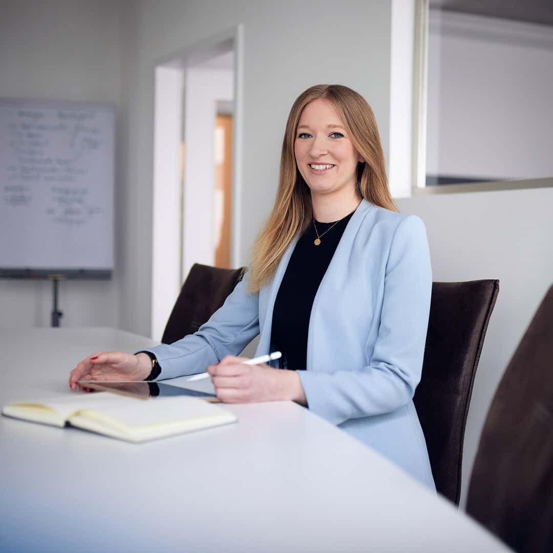 Eine Frau in einem blauen Blazer sitzt lächelnd an einem Tisch mit Notizbuch und Stift.