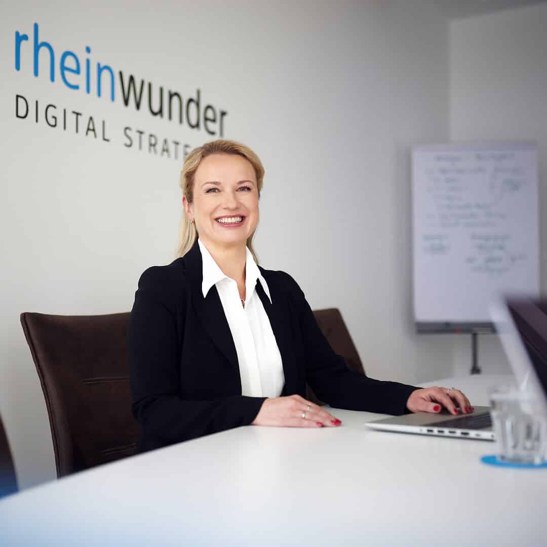 Eine lächelnde Frau im Anzug sitzt am Tisch mit Laptop, Flipchart im Hintergrund.