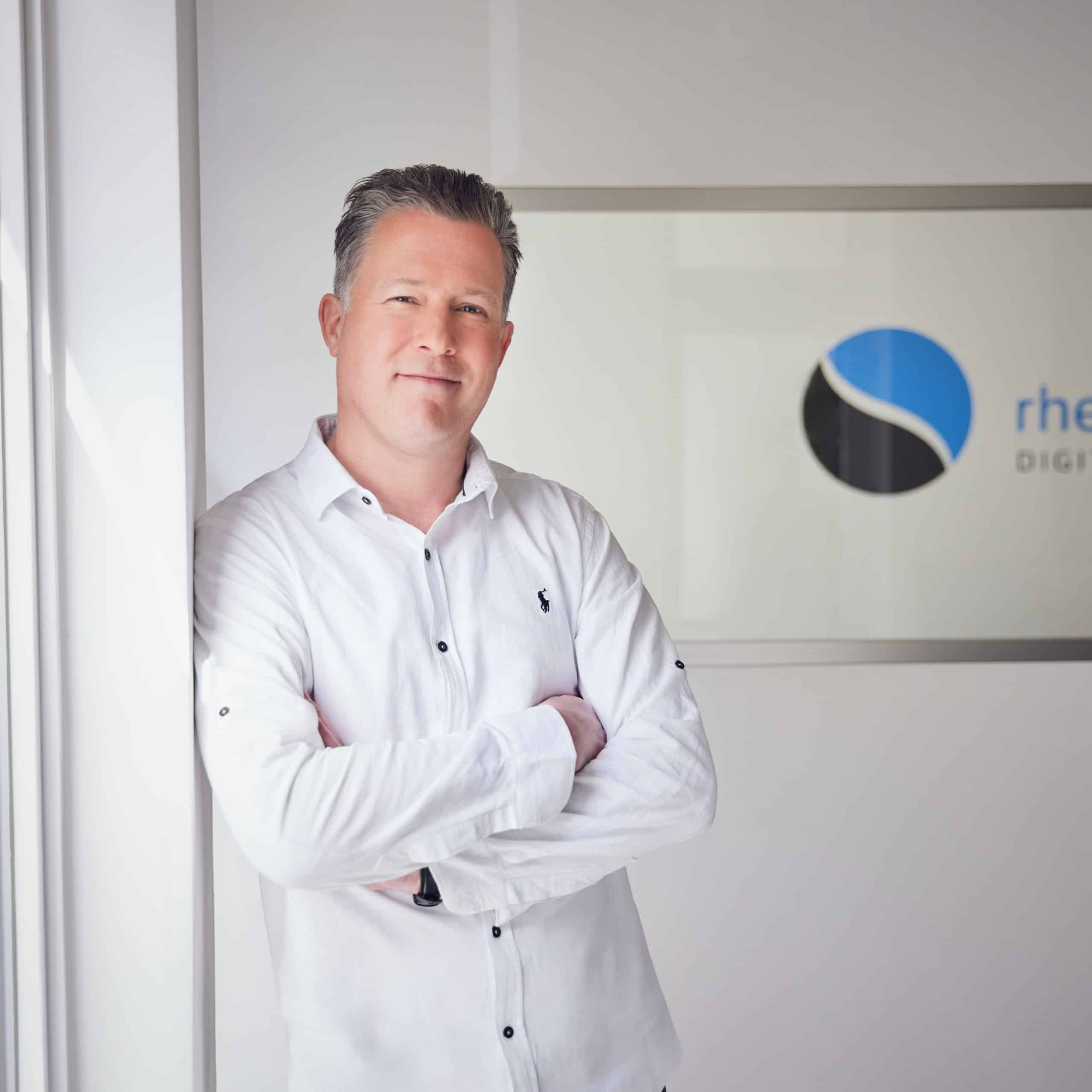 Ein Mann in weißem Hemd lehnt an einer Wand, neben ihm das Rheinwunder Logo