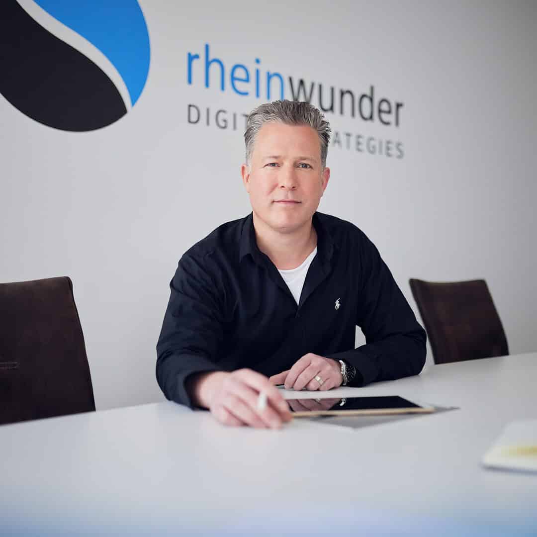 Ralph Grundmann sits at a table with a tablet, the Rheinwunder lettering on the wall behind him.