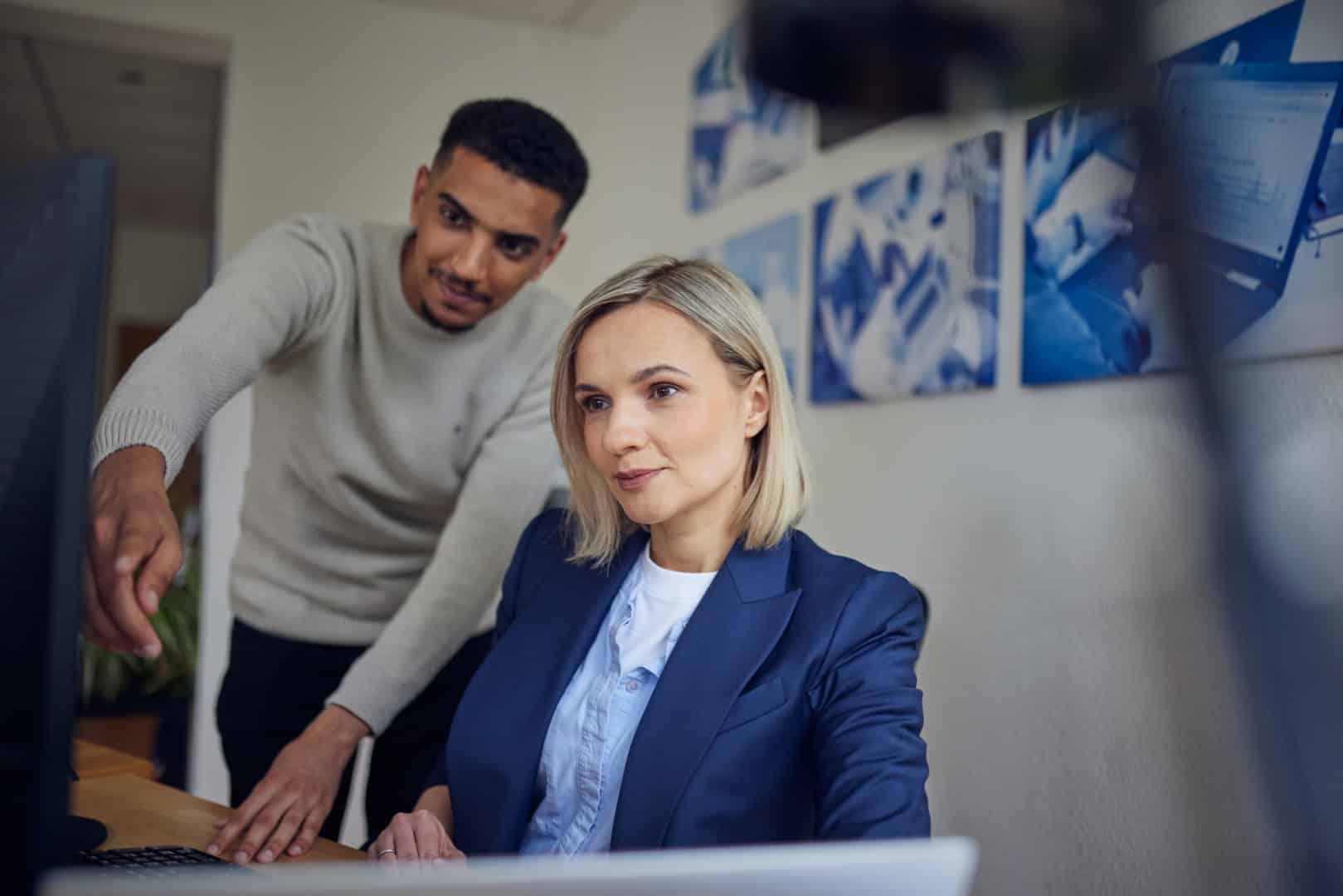 Mann zeigt auf Bildschirm, Frau im Anzug schaut konzentriert, Büro im Hintergrund.