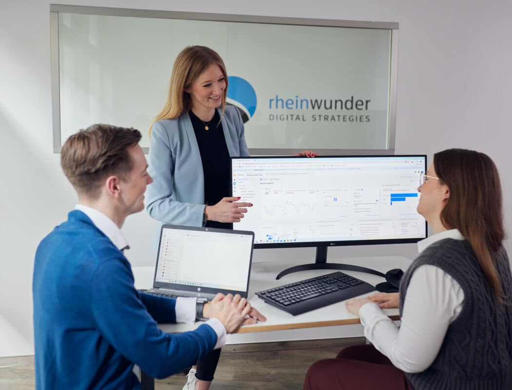Drei Personen in einem Büro, eine präsentiert Daten auf einem großen Monitor, zwei sitzen mit Laptops am Tisch.