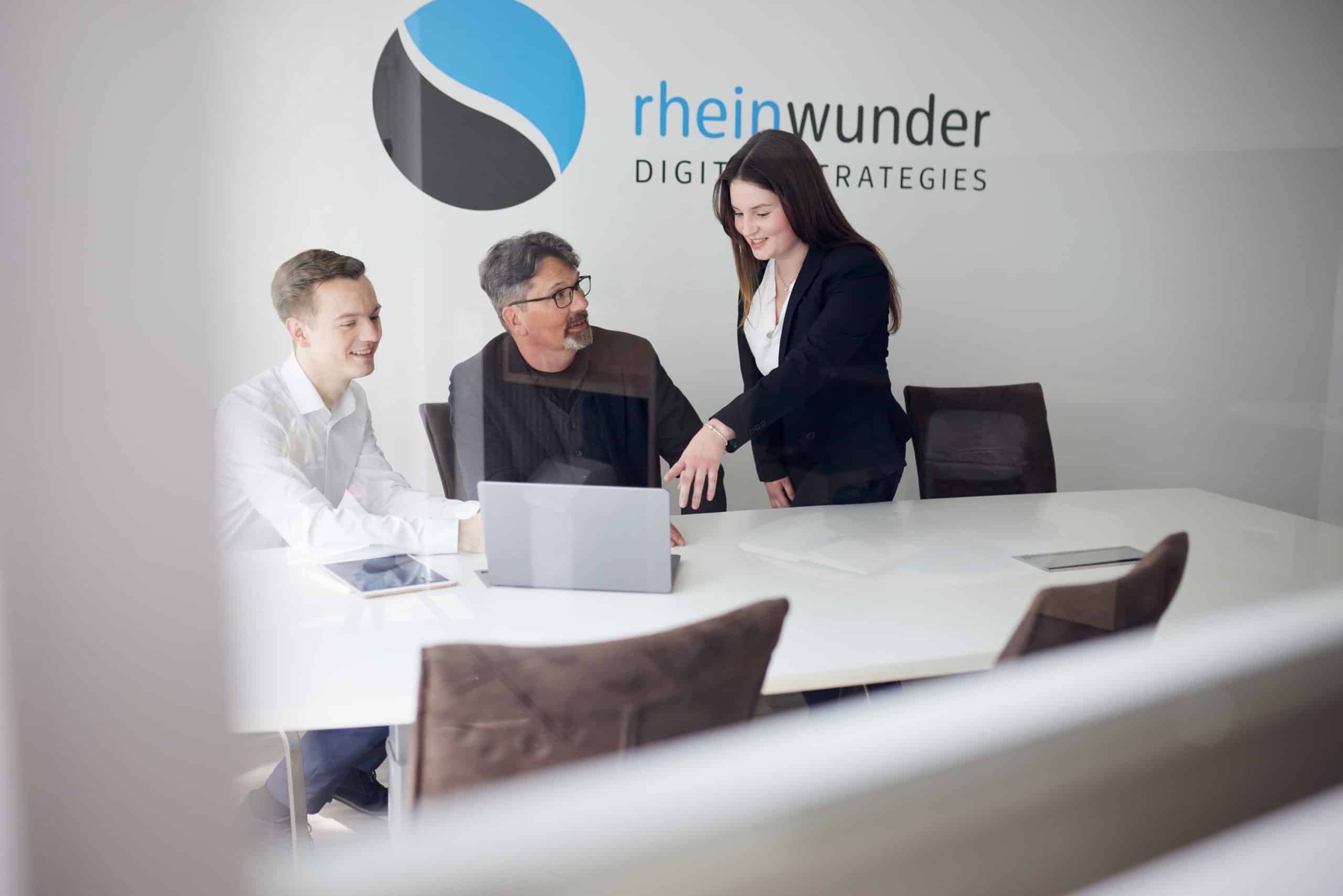 Drei Personen bei einem Meeting an einem Tisch mit Laptop, Logo von 'rheinwunder' im Hintergrund.