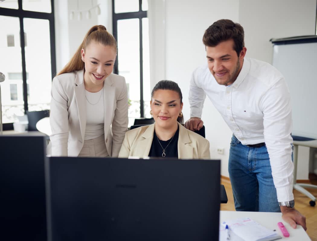 Drei Personen in Business-Kleidung schauen lächelnd auf einen Computerbildschirm in einem Büro.