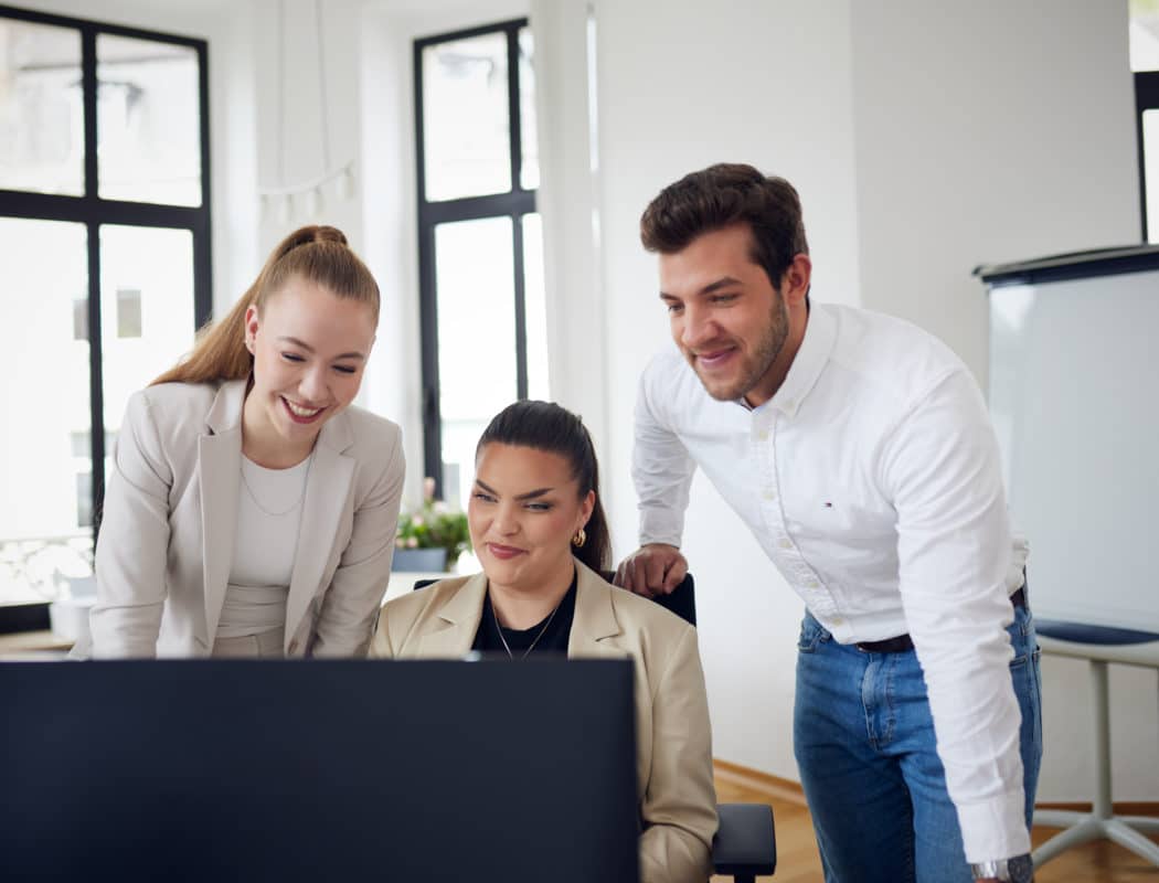 Drei Personen in Business-Kleidung lächeln und schauen auf einen Computerbildschirm in einem hellen Büro.