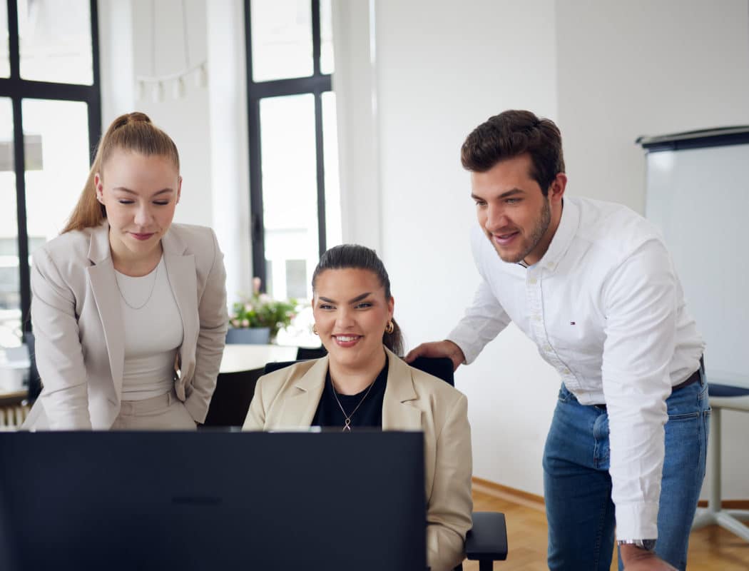 Drei Personen in Business-Kleidung lächeln vor einem Computerbildschirm in einem Büro.