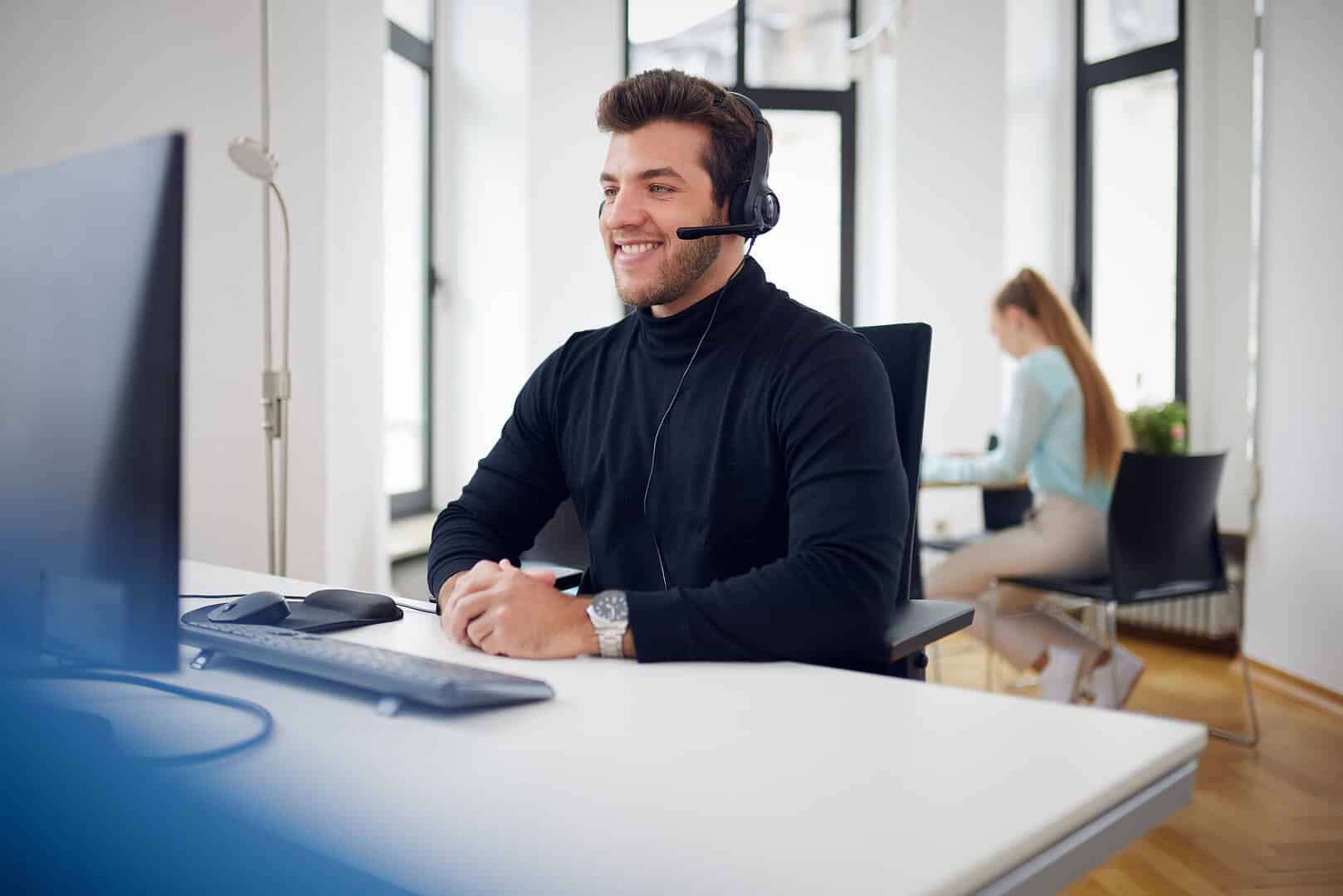 A man with a headset sits smiling at a desk, a woman works in the background.