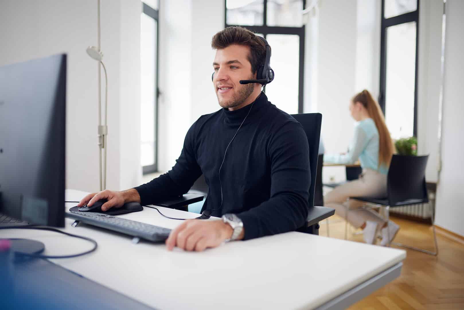 A man with a headset works at a computer, a woman sits at a table in the background.