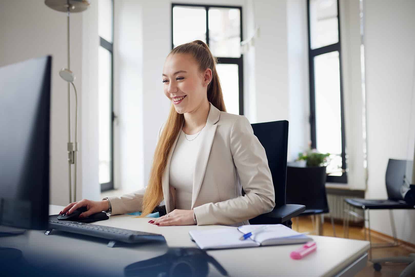 Eine Frau in einem Büro arbeitet lächelnd am Computer, ein Notizbuch und Stift liegen auf dem Tisch.