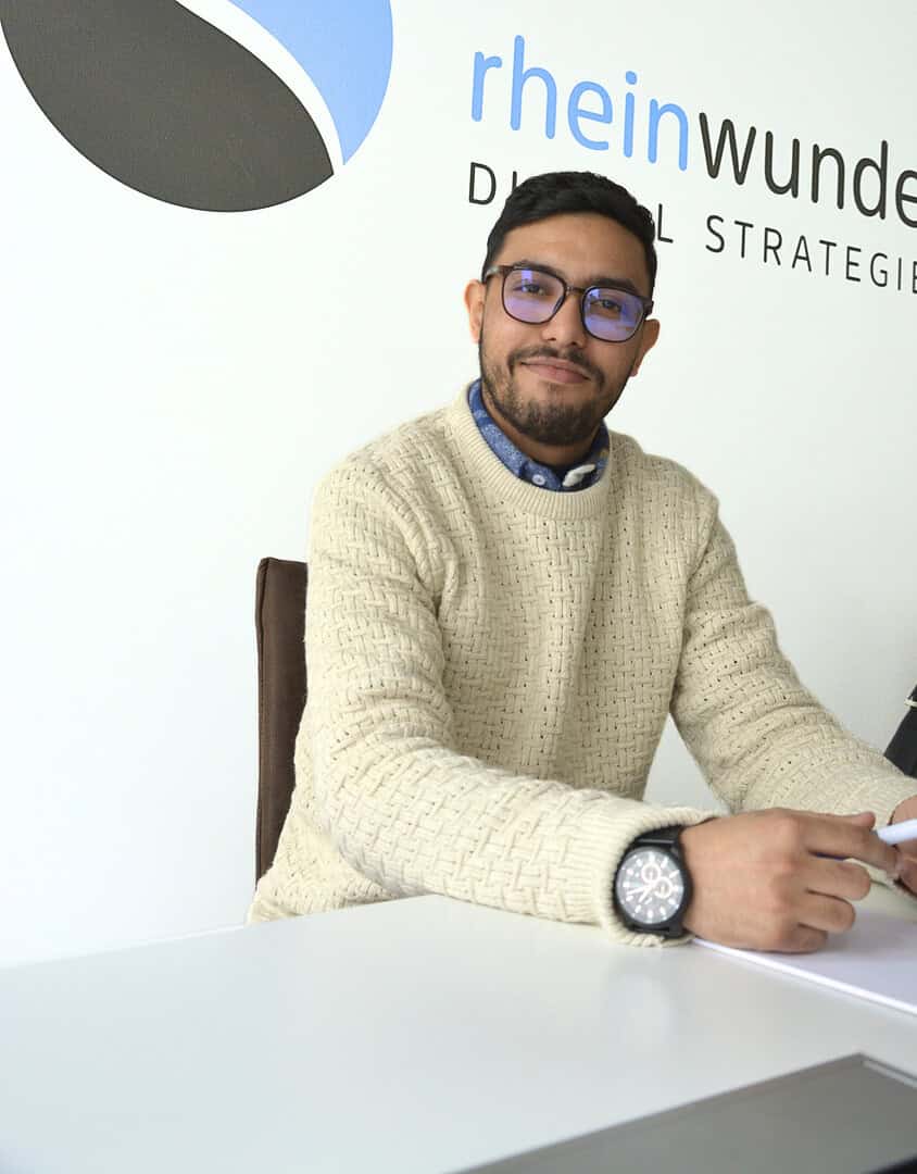 Eine Person sitzt lächelnd an einem Tisch vor einer Wand mit dem Logo rheinwunder.