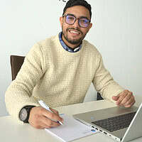 A man with glasses and a jumper writes at a table with laptop and notepad.